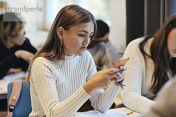 Teenager-Mädchen mit braunen Haaren  das ein Smartphone benutzt und neben einer Freundin am Schreibtisch im Klassenzimmer sitzt