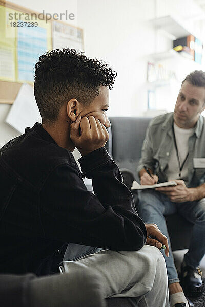 Depressiver Junge sitzt mit dem Kopf in der Hand bei einem männlichen Berater und diskutiert im Schulbüro