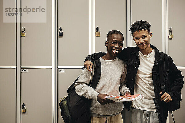 Porträt von lächelnden Teenagern  die Rucksäcke tragen  während sie vor einem Spind in der Schule stehen