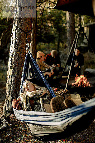 Junge Frau entspannt sich in einer Hängematte mit Freunden beim Zelten im Hintergrund