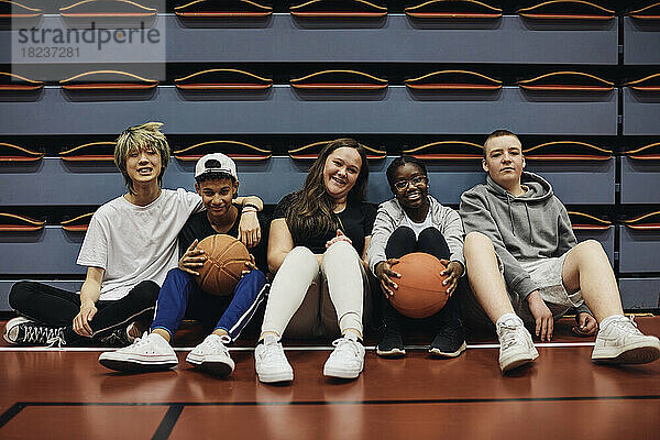 Porträt von lächelnden männlichen und weiblichen Freunden  die mit Basketbällen auf einem Sportplatz sitzen