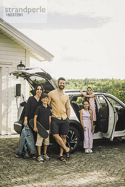 Porträt einer glücklichen Familie  die zusammen vor einem Elektroauto steht