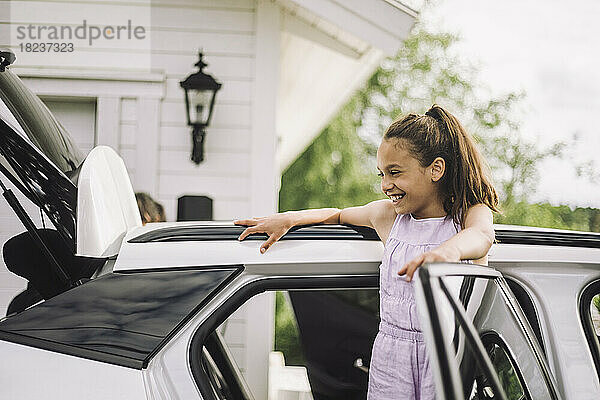Glückliches Mädchen im Grundschulalter lehnt sich an die Tür eines Elektroautos