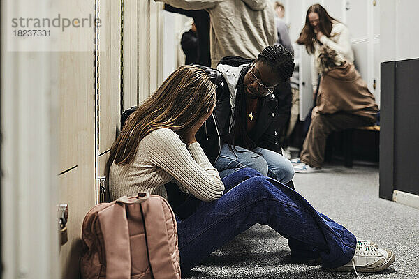 Teenager-Mädchen im Gespräch mit einer depressiven Freundin  die in einem Schulkorridor neben einem Rucksack sitzt