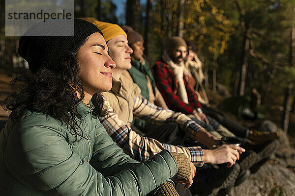 Seitenansicht einer lächelnden Frau mit geschlossenen Augen  die das Sonnenlicht genießt  während sie bei Freunden im Wald sitzt