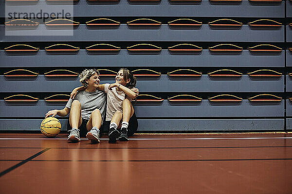 In voller Länge von lächelnden Mädchen sitzen mit Armen um einander auf Basketballplatz in der Schule