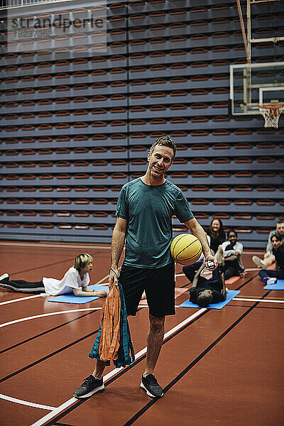 Ganzkörperporträt eines lächelnden männlichen Trainers  der mit einem Basketball gegen Schüler auf einem Sportplatz steht