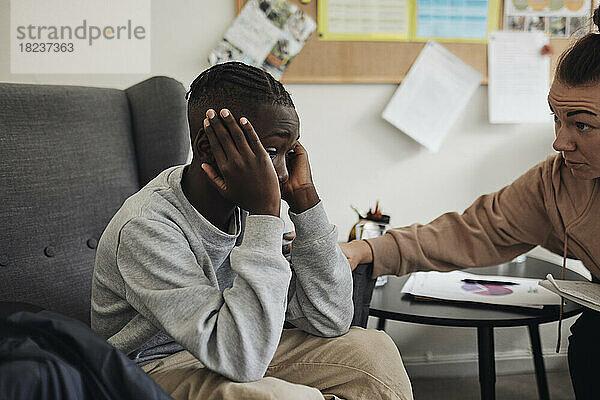 Nicht-binäre psychologische Fachkraft  die einen traurigen männlichen Studenten tröstet  der auf einem Stuhl im Büro sitzt