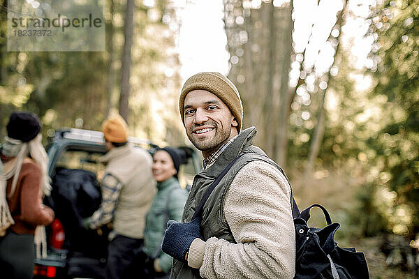 Seitenansicht eines lächelnden Mannes mit Rucksack und Freunden im Hintergrund