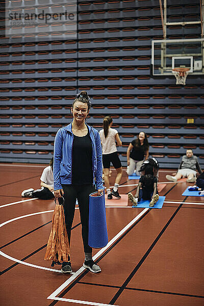Ganzes Porträt eines lächelnden weiblichen Trainers  der mit einer Übungsmatte auf einem Sportplatz steht