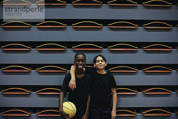 Glücklicher Jugendlicher mit Basketball  der neben einem männlichen Freund auf dem Sportplatz steht