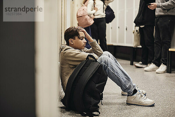 Seitenansicht eines traurigen Teenagers  der mit einem Rucksack im Schulflur sitzt