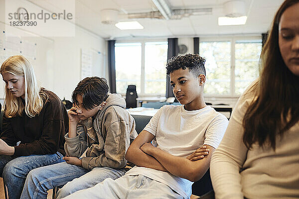 Nachdenklicher Teenager sitzt mit verschränkten Armen inmitten von Freunden im Klassenzimmer während einer Gruppentherapie