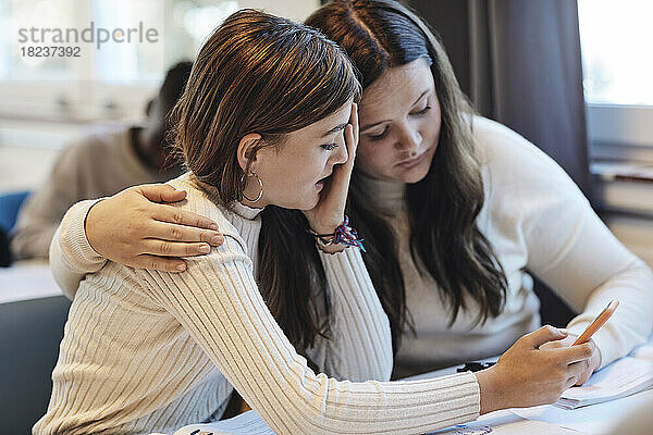Teenager-Mädchen mit Arm um Freundin teilen Smartphone mit ihr am Schreibtisch im Klassenzimmer
