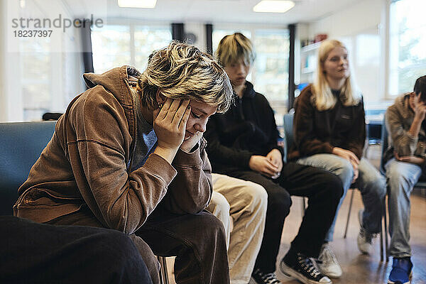 Trauriges Teenager-Mädchen mit dem Kopf in den Händen  das mit männlichen und weiblichen Freunden in einer Gruppentherapie sitzt