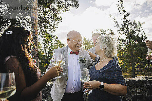 Lächelnde ältere Frau und Bräutigam mit Weingläsern  die sich bei der Hochzeit umarmen
