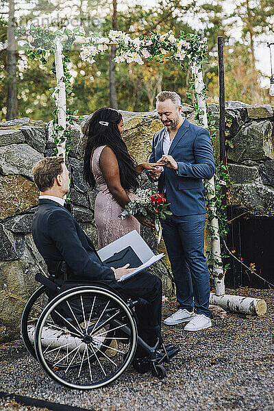 Lächelnder Bräutigam und Braut tauschen bei der Hochzeitszeremonie den Ring mit einem behinderten Pfarrer im Rollstuhl