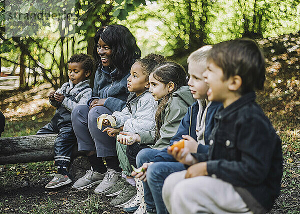Gemischtrassige Jungen und Mädchen essen Obst  während sie mit einer Lehrerin im Park sitzen