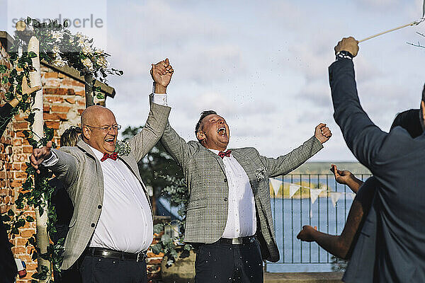 Glückliches  frisch verheiratetes schwules Paar  das sich an den Händen hält  während es seine Hochzeitszeremonie mit den Gästen genießt