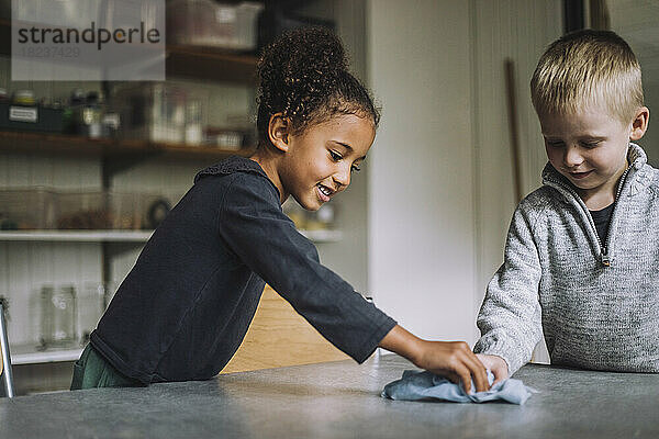 Lächelndes Mädchen und Junge säubern den Esstisch mit einer Serviette in einer Kindertagesstätte