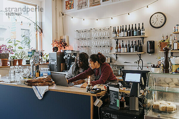 Lächelnde junge Besitzerin  die mit einem Kollegen an der Kasse eines Cafés über einen Laptop diskutiert