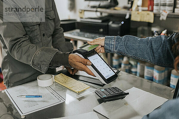 Männlicher Schneider hält ein Kreditkartenlesegerät in der Hand  während eine Kundin in einer Werkstatt kontaktlos bezahlt