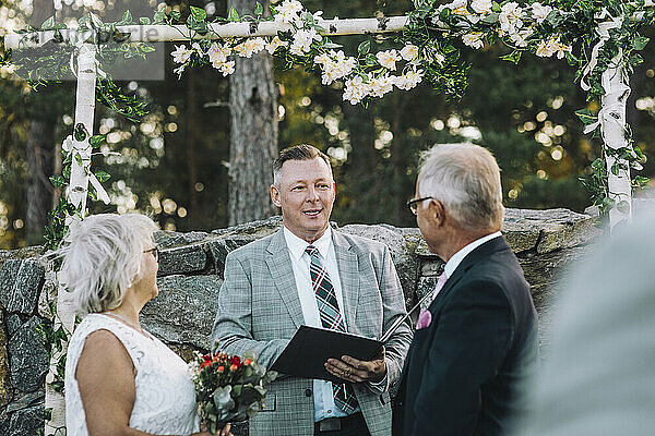 Älteres Paar tauscht Gelübde aus  während es auf den Minister blickt  der während der Hochzeit an der Wand mit Blumenschmuck steht