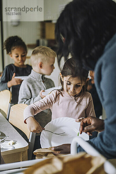 Mädchen wirft Essensreste in Papiertüte  die von Lehrerin im Kindergarten gehalten wird