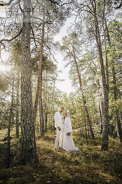 Mid Erwachsenen Braut hält Bouquet Blick auf Bräutigam stehend inmitten von Bäumen im Wald