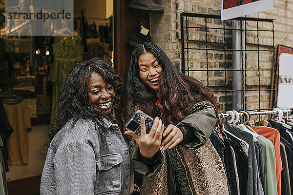 Glückliche Freundinnen  die ein Selfie mit ihrem Smartphone machen  während sie sich vor einem Bekleidungsgeschäft vergnügen