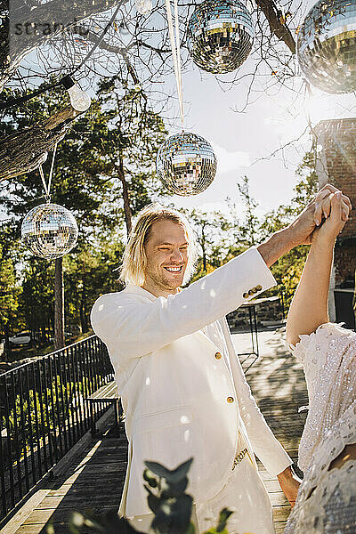 Glücklicher Bräutigam hält die Hand der Braut und tanzt unter Discokugeln an einem sonnigen Tag