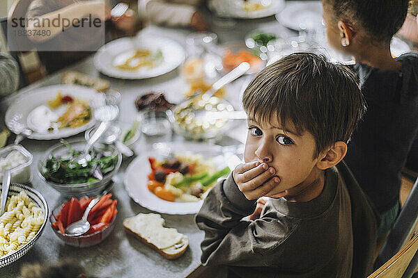 Porträt eines Jungen  der den Mund mit der Hand bedeckt  während das Frühstück auf dem Tisch einer Kindertagesstätte steht