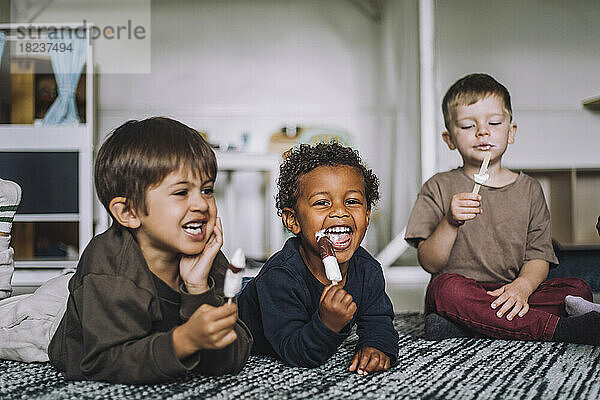 Fröhliche männliche Studenten essen Eis auf dem Teppich in der Kindertagesstätte