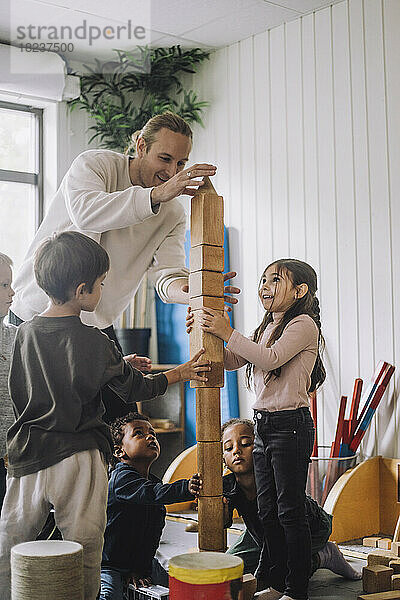 Männlicher Lehrer  der Kinder beim Stapeln von Holzklötzen im Kindergarten anleitet