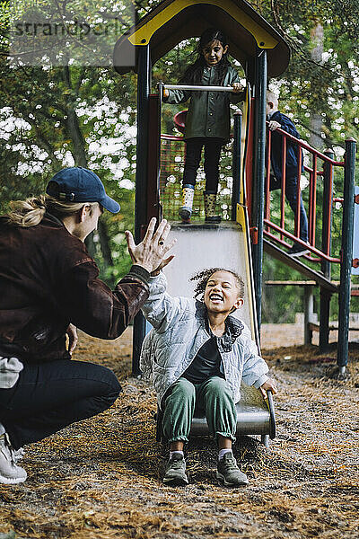 Fröhliches Mädchen gibt männlichem Lehrer High-Five  während sie auf dem Spielplatz auf der Rutsche spielt