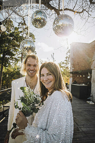 Glückliche Braut mit Blumenstrauß und Bräutigam unter Discokugeln an einem sonnigen Tag