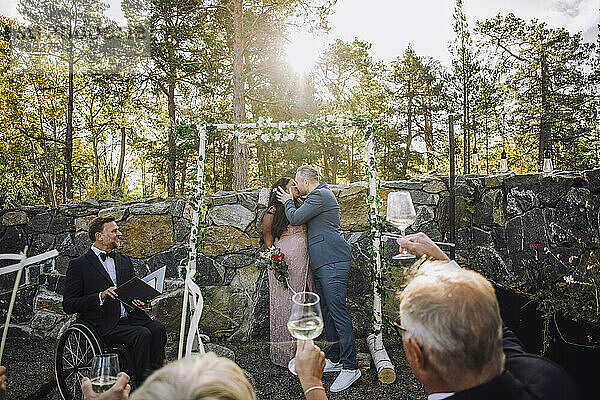 Neuvermähltes Paar küsst sich vor den Gästen  die bei der Hochzeitszeremonie Trinksprüche ausbringen  und einem Pfarrer mit Behinderung