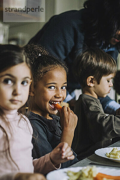 Glückliches Mädchen isst Karotte zum Frühstück mit Klassenkameraden im Kindergarten