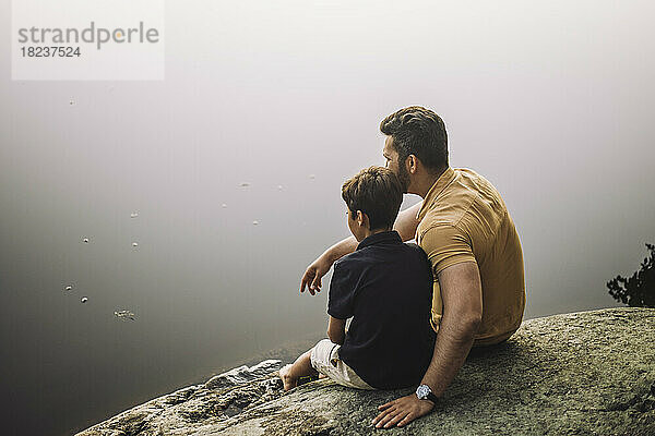 Vater sitzt mit Sohn auf einem Felsen am See