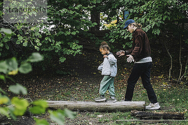 Seitenansicht eines Mädchens  das auf einem Baumstamm balanciert  während ein männlicher Lehrer sie im Park führt