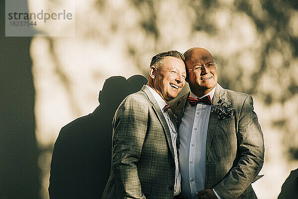 Glückliche gut gekleidete Homosexuell Männer stehen gegen die Wand während der Hochzeit am sonnigen Tag