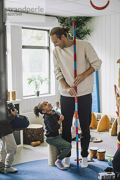 Lächelnder männlicher Lehrer  der eine Schülerin ansieht  die in einer Kindertagesstätte auf einer Trommel sitzt