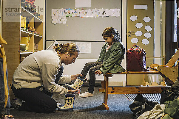 Männlicher Lehrer hilft Mädchen beim Anziehen von Gummistiefeln  während sie in der Kindertagesstätte sitzen