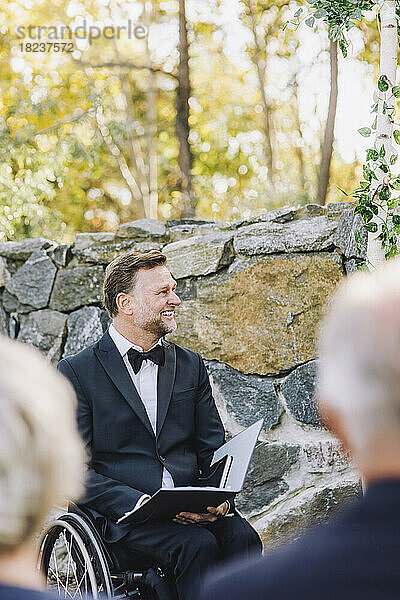 Glücklicher Pfarrer im Rollstuhl mit Behinderung  der während der Hochzeit ein Buch an die Wand hält