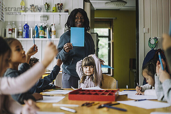 Fröhliche Lehrerin zeigt Schülern farbiges Papier  während sie sie in der Kindertagesstätte unterrichtet