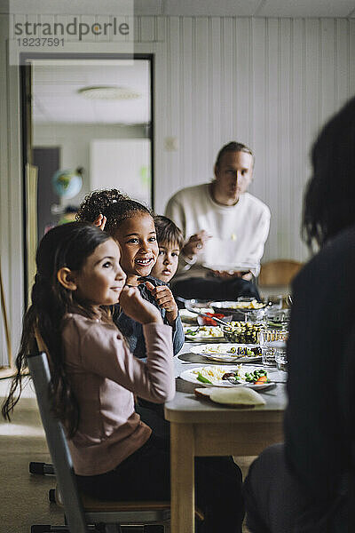 Lächelndes Mädchen im Gespräch mit der Lehrerin und Klassenkameraden beim Frühstück im Kindergarten