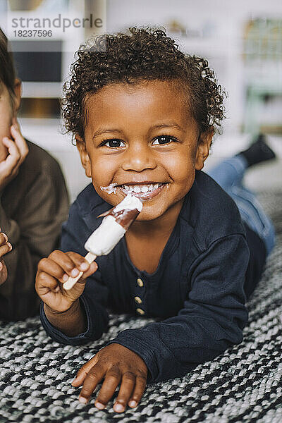 Porträt eines glücklichen Jungen  der von einem männlichen Mitschüler auf dem Teppich liegend ein Eis isst  im Kindergarten