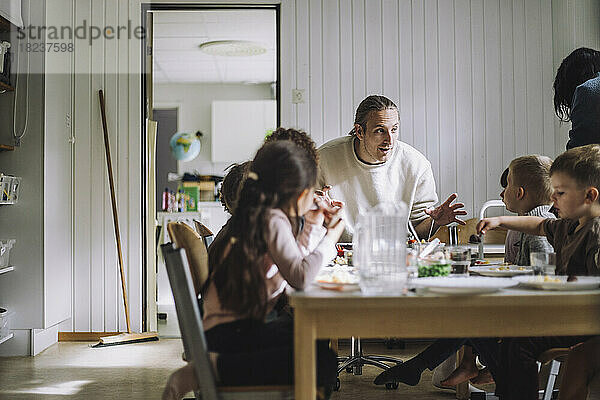 Männlicher Lehrer im Gespräch mit Schülern beim Frühstück am Tisch in einer Kindertagesstätte