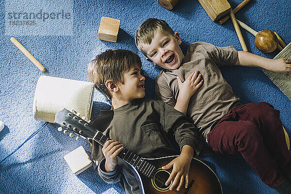 Fröhlicher Junge im Liegen mit einem Mitschüler  der eine Gitarre hält  im Kindergarten