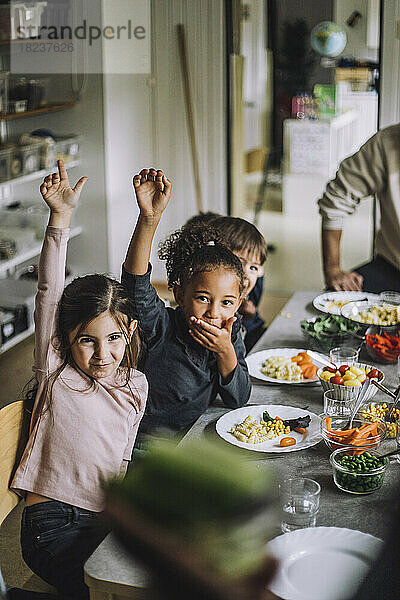 Glückliche Mädchen und Jungen mit erhobenen Händen am Esstisch beim Frühstück im Kindergarten